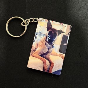 May include: A rectangular keychain featuring a color photograph of a dog. The dog has brown and black fur and is wearing a collar. The keychain has a silver-colored metal ring and chain.