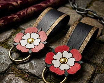 Pode incluir: Duas pulseiras de couro preto com acabamentos dourados e um design de rosa Tudor vermelha, branca e verde. Cada pulseira tem um anel dourado. Apresentadas numa superfície de pedra.