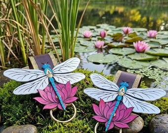 Pode incluir: Dois porta-guardanapos decorativos em forma de libélula. Cada um apresenta asas brancas e pretas, um corpo azul e uma base de flor de lótus rosa. Os porta-guardanapos têm um anel dourado e uma faixa de couro castanho.
