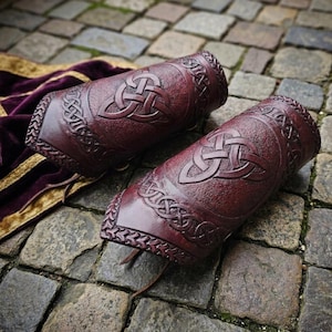 May include: Pair of burgundy leather arm guards with Celtic knot designs. The arm guards feature embossed knotwork and decorative borders. A leather strap is visible on each guard. The background is a stone surface.