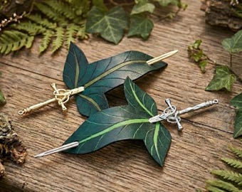Pode incluir: Dois ganchos de cabelo em forma de folhas verde-escuras com nervuras proeminentes. Um gancho apresenta um alfinete de metal dourado em forma de espada, enquanto o outro apresenta um alfinete de metal prateado. Os alfinetes atravessam as folhas.