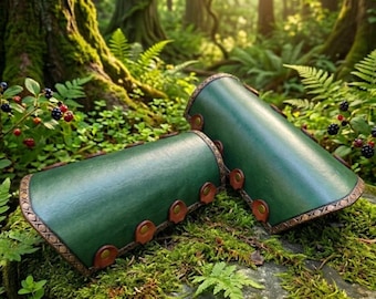Pode incluir: Um par de braçadeiras de couro verde-floresta com acabamento em couro castanho nas bordas, fixadas com rebites metálicos. Os protetores de braço cilíndricos têm um acabamento liso e estão posicionados horizontalmente sobre musgo.