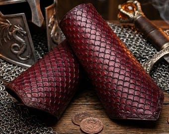 Pode incluir: Protetores de braço de couro bordô com padrão de escamas, exibidos em uma superfície de madeira. Um capacete medieval, cota de malha, moedas e uma espada ao fundo, sugerindo um tema de fantasia ou histórico.