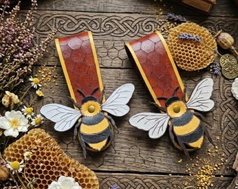 Pode incluir: Dois marcadores de livros de couro com enfeites de abelha. Os marcadores de livros têm um padrão de favo de mel marrom e uma borda amarela. Cada abelha tem um corpo amarelo e preto com asas brancas. O fundo é uma superfície de madeira com detalhes florais e de favo de mel.