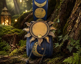 Pode incluir: Uma bolsa de couro azul-escura com um emblema de sol dourado na aba frontal e um fecho de pressão em latão. A alça vertical exibe três fases da lua em dourado, enquanto a seção inferior mostra uma lua crescente dourada e estrelas.