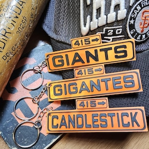 May include: Orange and black keychains with the words "GIANTS", "GIGANTES", and "CANDLESTICK" are displayed. A baseball bat and a San Francisco Giants baseball cap are also in the image.