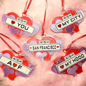 May include: Heart-shaped ornaments in red and purple with white license plate-style signs. The signs read "YOU", "MY CITY", "A ❤️ F", "SAN FRANCISCO", and "MY HOOD". Each ornament has a red ribbon for hanging.