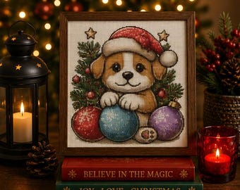 May include: A framed cross-stitch embroidery featuring a puppy wearing a red Santa hat, positioned in front of a pine tree with red, blue, and purple ornaments. The artwork rests on two stacked books with the text Believe in the Magic and Joy Love Christmas.