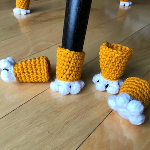 May include: Close-up of five crocheted chair leg covers. The covers are yellow with white pom-poms. The covers are on a wooden floor.