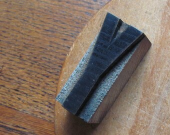 May include: A wooden printing block with the letter "Y" carved into it. The block is dark brown and has a rough texture. It is sitting on a light brown wooden surface.