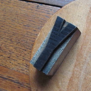 May include: A wooden printing block with the letter "Y" carved into it. The block is dark brown and has a rough texture. It is sitting on a light brown wooden surface.