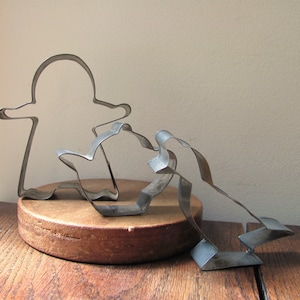 以下が含まれることがあります： 木製の表面に置かれた、ヴィンテージの金属製のクッキー型3つ。クッキー型は、人、鳥、ティーポットの形をしています。