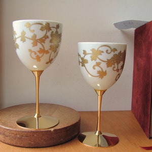 Puede incluir: Dos copas de vino blanco con diseños florales de pan de oro sobre una superficie de madera. Las copas tienen un tallo y una base dorados.