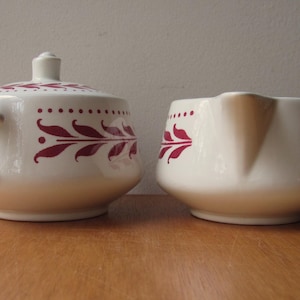 May include: A white ceramic sugar bowl and creamer set with a red leaf design. The sugar bowl has a lid.