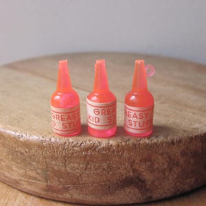 May include: Three small, translucent, orange-red plastic bottles with white labels. The labels read "GREASY STUFF" and "GREAT KID STUFF". The bottles are arranged on a round, wooden surface.