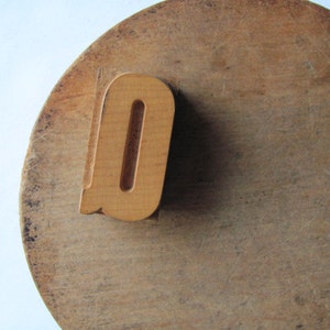 May include: A wooden letter "O" stamp sits on a round, well-worn wooden surface. The stamp is a light brown color, and the wooden surface has a darker, aged appearance with visible grain and wear marks. The background is white.