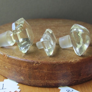 May include: Three clear glass bottle stoppers with different shapes. The stoppers are on a wooden surface.
