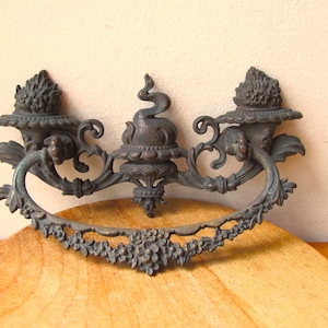 May include: An ornate, antique-style bronze drawer pull with a decorative, floral design. The pull features a central, raised, stylized serpent design and two smaller, decorative finials on either side.