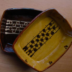Può includere: Due piatti da portata rettangolari in ceramica. Uno è marrone scuro con un disegno bianco e crema. L'altro è giallo con un motivo marrone. Entrambi hanno un bordo marrone rossiccio e sono su una superficie marrone chiaro.