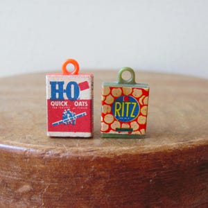 May include: Two miniature food box charms. One is a red, white, and blue box of "HO Quick Oats" with an orange loop. The other is a green and red box of "RITZ" crackers with a green loop. Both are on a wooden surface.