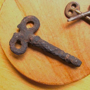 May include: A heavily rusted, dark brown metal winding key with a figure-eight shaped handle and a straight, textured shaft. A second, smoother, brass-colored metal key with a similar figure-eight handle is partially visible in the upper corner.
