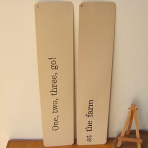 May include: Two rectangular cards with rounded corners. One card reads "One, two, three, go!" and the other reads "at the farm" in black text. The cards are a light tan color. A small wooden easel is in the background.