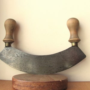 Może przedstawiać: Zabytkowy nóż mezzaluna z zakrzywionym, postarzanym ostrzem ze stali i drewnianymi uchwytami. Ostrze jest oznaczone tekstem i numerem 11. Nóż jest prezentowany na małej, okrągłej drewnianej podstawie.