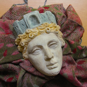 May include: A plaster sculpture of a classical face with a crown, set on patterned fabric. The face is off-white with closed eyes and curly blonde hair. The crown is light blue with a castle design.