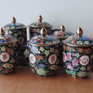 Pode incluir: Seis chávenas de chá pretas com tampas, cada uma decorada com padrões florais coloridos e detalhes dourados. As tampas têm botões dourados e as chávenas têm alças. As chávenas são decoradas com caracteres chineses.