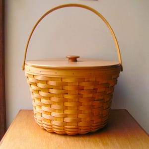 Pode incluir: Uma cesta tecida castanha clara com tampa e alça de madeira. A cesta é redonda com uma alça curva e um pequeno botão de madeira na tampa. A cesta está sobre uma superfície de madeira.