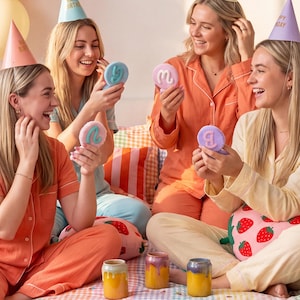 May include: Four individuals in pajamas and party hats sit on a bed with a colorful plaid comforter, holding small round cookies with icing letters. The room is decorated with orange and yellow balloons and a Happy Birthday banner.
