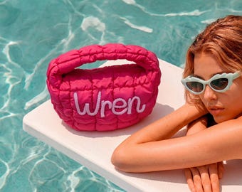 Puede incluir: Un bolso de mano acolchado de color rosa brillante con un asa redondeada y el nombre Wren bordado en letra blanca en la parte delantera. El bolso tiene una textura suave y esponjosa y está colocado sobre una superficie blanca junto a unas gafas de sol verde claro.