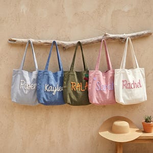 May include: Five canvas tote bags in various colors: light blue, denim, olive green, pink, and white. Each bag has a name embroidered on it: Piper, Kayle, Rala, Sarah, and Rachel. The bags are hanging on a wooden branch.