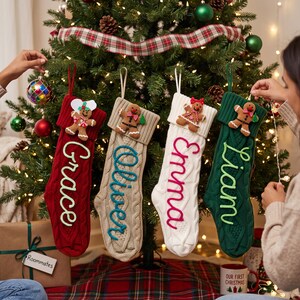 Può includere: Quattro calze natalizie in maglia a trecce nei colori rosso, beige, bianco e verde scuro, ciascuna con un omino di pan di zenzero e un nome ricamato in corsivo: Grace, Oliver, Emma e Liam, appese a un albero.