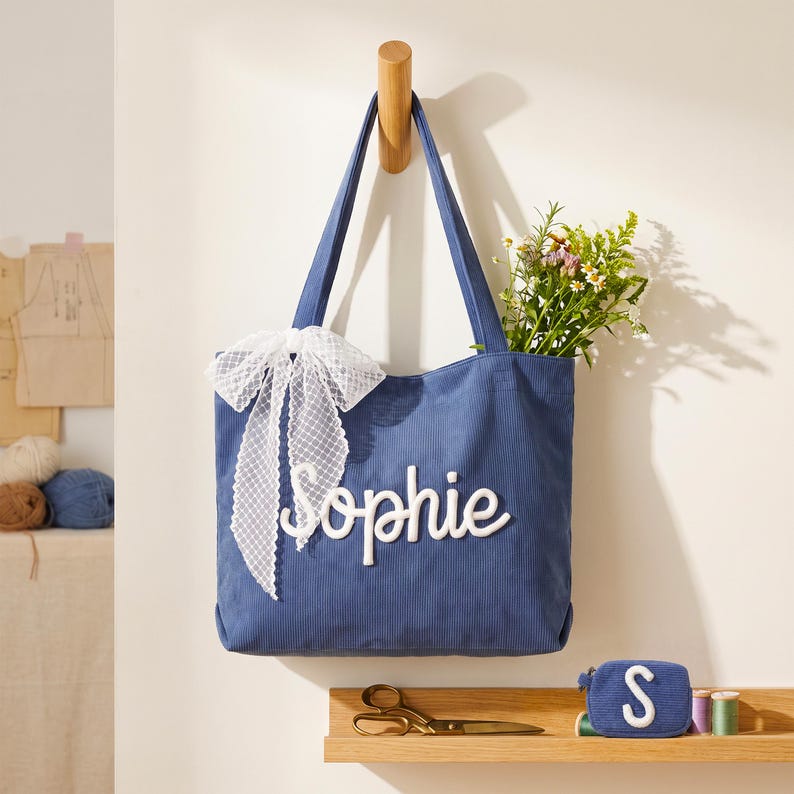 May include: A blue corduroy tote bag featuring the name Sophie in white raised lettering and a white lace ribbon tied to the side. The bag contains a bouquet of wildflowers and is displayed alongside a small matching blue coin purse with an S.