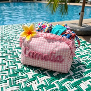 Puede incluir: Un neceser de tela con estampado de cuadros vichy en rosa y blanco, con un borde superior con volantes y el nombre Camelia bordado en hilo rosa en el frente. Una flor de plumeria amarilla y blanca est&aacute; sujeta al lateral, con telas estampadas.