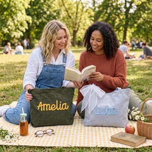 May include: Two rectangular tote bags made of fabric. One bag is olive green with the name Amelia in yellow script lettering. The other bag is light gray with the name Luna in light blue script lettering and a white polka dot ribbon bow attached.