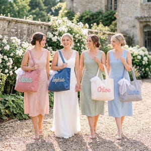 May include: Four tote bags in pink, blue, cream, and light blue, each featuring a name embroidered in cursive script: Olivia, Sophie, Chloe, and Luna. Each bag includes a white ribbon tied to one handle, showcasing a simple, functional design.