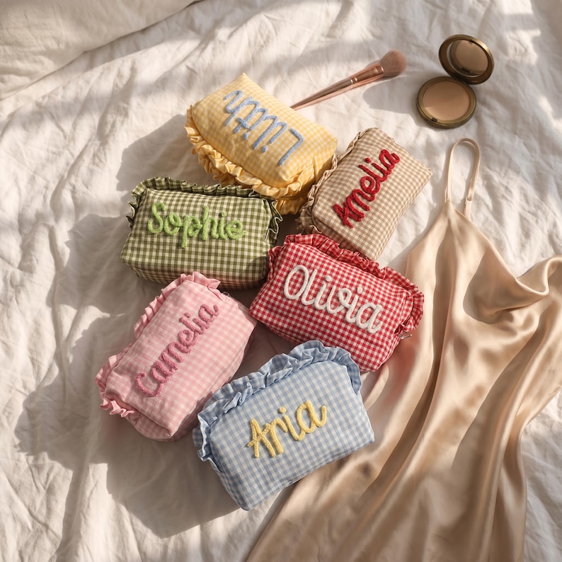 Puede incluir: Seis estuches rectangulares de tela con bordes de volantes y patrones de cuadros vichy en amarillo, verde, rosa, rojo, beige y azul. Cada estuche tiene un nombre bordado en letra cursiva: Lilith, Sophie, Camelia, Amelia, Olivia y Aria.