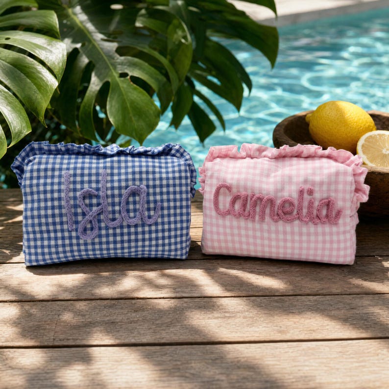 Puede incluir: Dos estuches rectangulares de tela con bordes de volantes y estampado de cuadros vichy. Uno es azul y blanco con el nombre Isla bordado en hilo morado. El otro es rosa y blanco con el nombre Camelia bordado en hilo rosa.