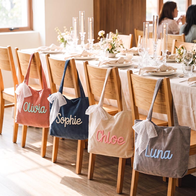 May include: Four rectangular tote bags in dusty rose, navy blue, beige, and gray, each featuring a white lace bow on the strap and a name embroidered in cursive script: Olivia, Sophie, Chloe, and Luna. The bags are hanging on wooden chairs.