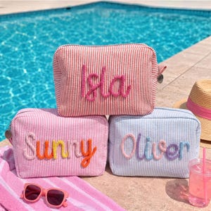 May include: Three rectangular zipped pouches made of seersucker fabric with thin vertical stripes. The top pouch is red and white with the name Isla in pink yarn. The bottom left is pink and white with Sunny in multi-coloured yarn. The bottom right is blue and white with Oliver in multi-coloured yarn.