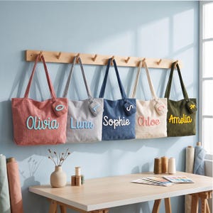 May include: Five corduroy tote bags in pink, gray, blue, cream, and olive green hanging on a wooden wall rack. Each bag features a personalized name in raised script lettering and a small matching coin pouch with a single embroidered letter.