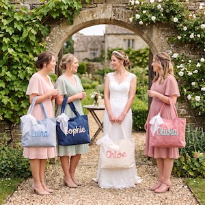 May include: Four rectangular canvas tote bags, each featuring a white lace ribbon tied to one handle and a name embroidered in cursive script on the front. The bags are light blue with Luna, dark blue with Sophie, white with Chloe, and pink with Olivia.