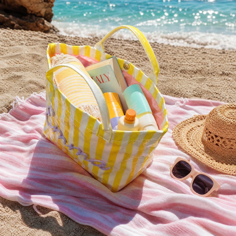 May include: A yellow and white striped tote bag with two handles, containing a book with the word Italy visible on the cover, a yellow bottle, and a light blue bottle. The bag features purple embroidered script text on the exterior side.