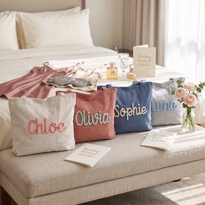 May include: Four canvas tote bags in cream, dusty rose, blue, and gray, each featuring a name in raised script lettering: Chloe, Olivia, Sophie, and Luna. The bags are arranged on a bench with wedding weekend schedule cards and perfume bottles.
