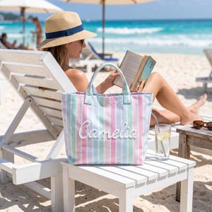 May include: A tote bag featuring vertical stripes in pastel shades of pink, blue, and green. The bag has two top handles and the name "Camelia" is embroidered in white script lettering across the center of the front panel of the bag.