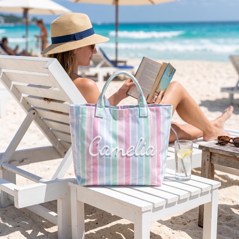 May include: A tote bag featuring vertical stripes in pastel shades of pink, blue, and green. The bag has two top handles and the name "Camelia" is embroidered in white script lettering across the center of the front panel of the bag.