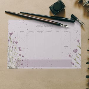 Puede incluir: Un planificador semanal con un diseño floral y las palabras "Weekly Planner" en la parte superior. El planificador tiene espacios para cada día de la semana, con flores de acuarela y plumas estilográficas.
