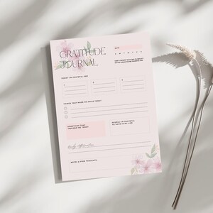 Puede incluir: Un diario de gratitud rosa con detalles florales y la frase "Gratitude Journal". Incluye indicaciones para la reflexión diaria, como "Hoy estoy agradecido por" y "Cosas que me hicieron sonreír hoy".